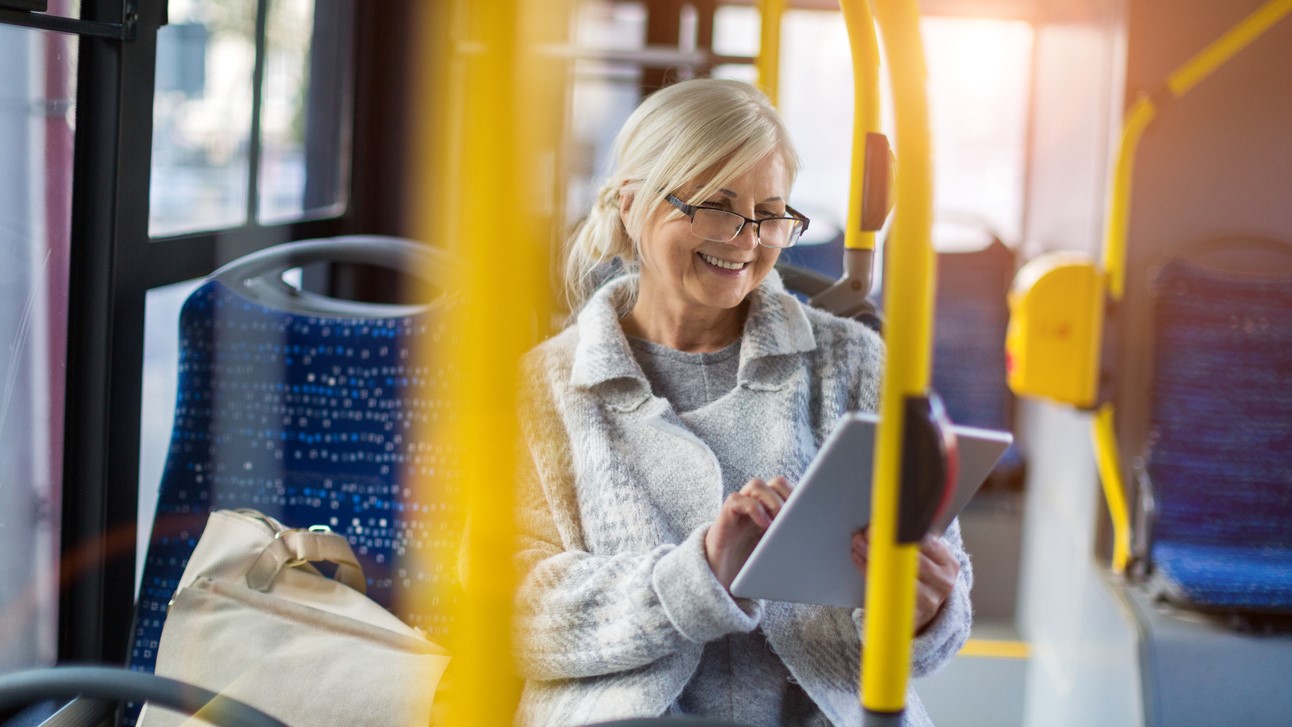Ældre kvinde med briller i en bus, smiler og kigger på iPad