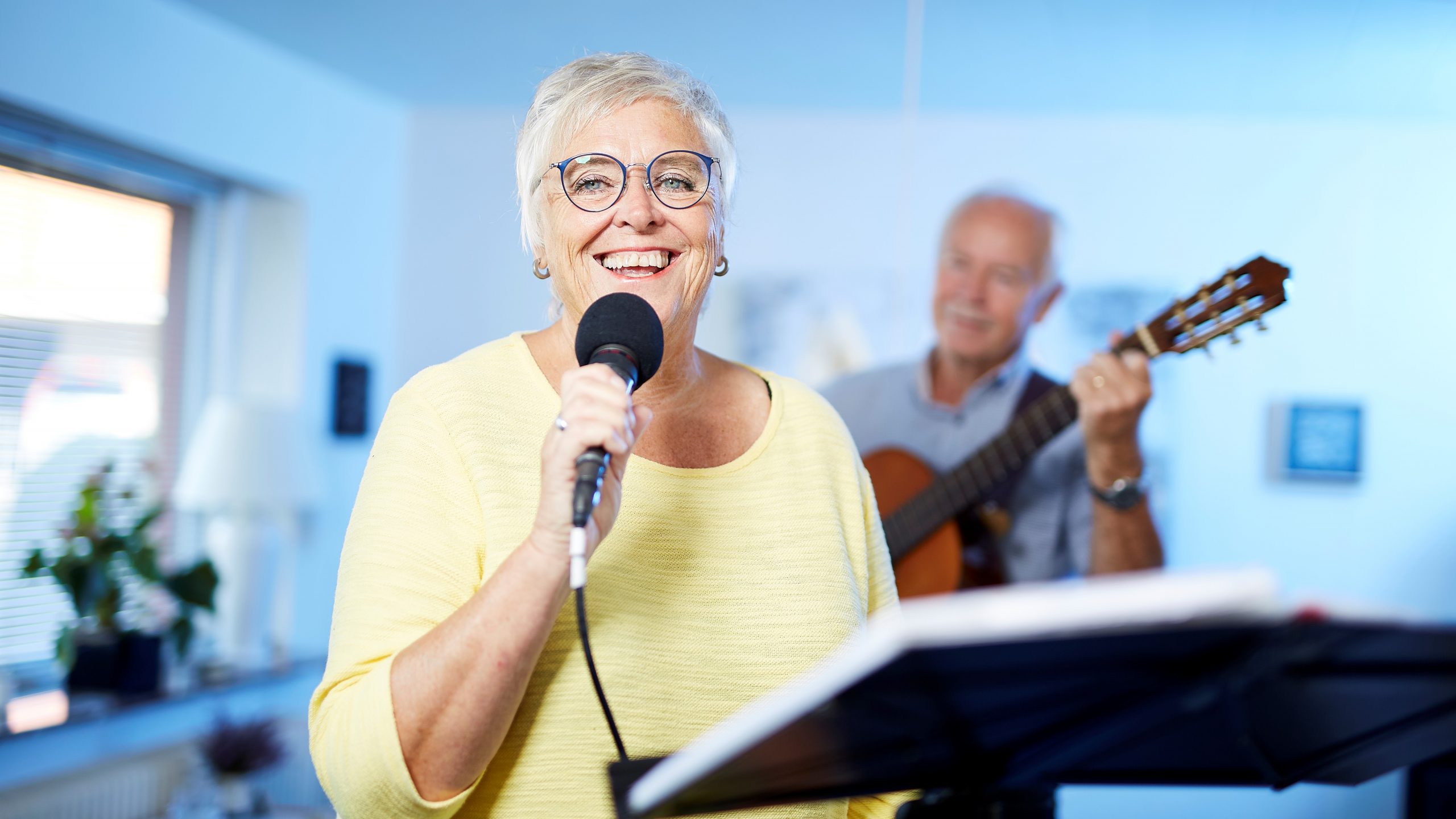 Smilende senior Henny med briller holder mirkrofon i hånden og i baggrunden ses en musiker