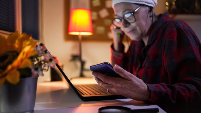 Senior kvinde med briller ser smilende på telefon, hun sidder ved et bord med computer og lup